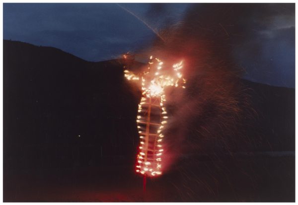 A fire effigy in the shape of a human figure with hands raised to the sides burns brightly against a hilly landscape at dusk