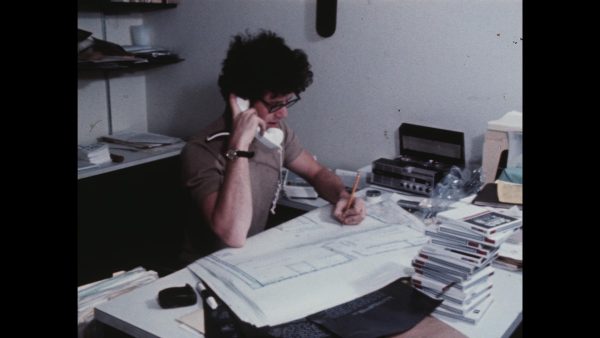A person sitting at a messy desk holds a white phone receiver to their ear as they write something down on a building blueprint with a pencil