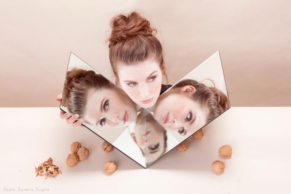 A person poses between two square mirrors that they hold in their hands to duplicate their head three times