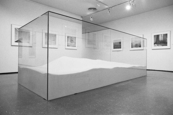 A large, clear, rectangular box filled with a sand-like material in the middle of a gallery space with pictures hanging on the walls