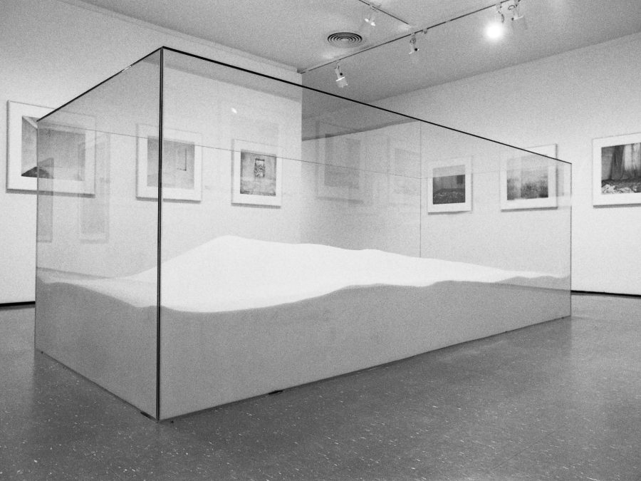 A large, clear, rectangular box filled with a sand-like material in the middle of a gallery space with pictures hanging on the walls
