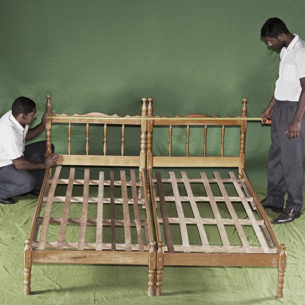 Two people wearing white shirts and black pants stand on opposite sides of a wooden bed frame in a green room.
