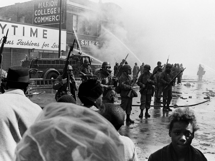 Archival black and white image of a group of people holding guns in gas masks. In front of them a small Black child looks directly at the camera while standing near other people.