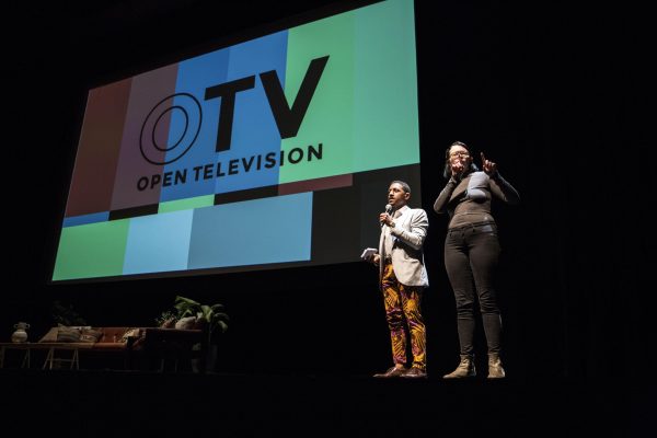 Two people on a stage with a large projection of text reading OTV Open Television behind them to the left