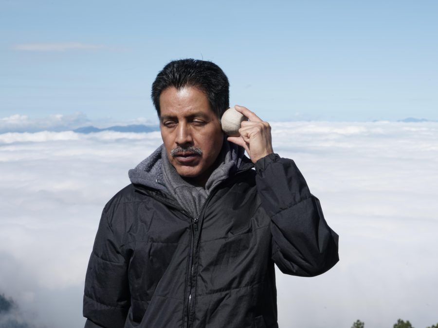 A man with medium-toned skin closes his eyes and holds a round, rock-like object against his temple. Behind him is a low cloud bank, indicating that he is standing at a very high elevation.