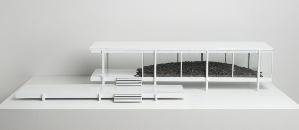 A model of a minimal, white building with floor-to-ceiling windows reveals an interior that holds a mound of dark, organic material.