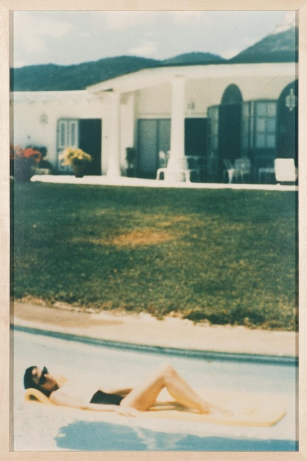 In what appears to be an aged photograph, a dark-haired person reclines on a lounge pool float in front of a large yard and house.