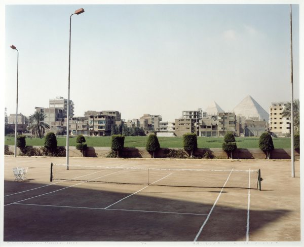 In a complex but quiet scene, a tennis court occupies the foreground. Behind the court are a line of modernist buildings, with the Egyptian pyramids looming in the distance.