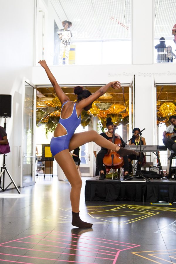 A dancer in a purple leotard kicks their leg up as their raise both hands in a bright space with a band behind them