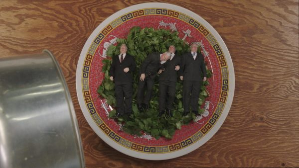 An ornately decorated plate holds a hearty serving of green herbs. Atop the herbs, four gray-haired men in suits lay side-by-side.