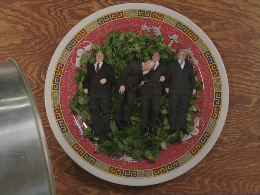 An ornately decorated plate holds a hearty serving of green herbs. Atop the herbs, four gray-haired men in suits lay side-by-side.