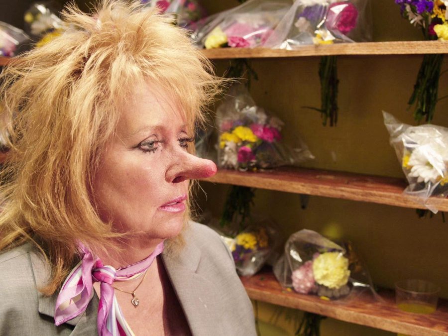 A woman with disheveled blonde hair and a comically long, pink-tipped nose is seen in front of wooden shelves of flower bouquets.