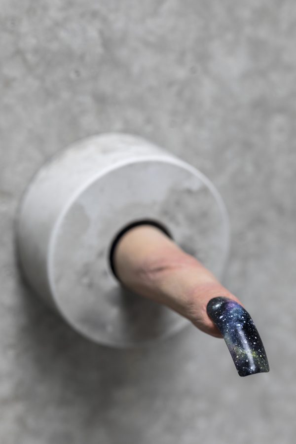 A light-skinned finger ending in a long fingernail extends out from a gray wall. The fingernail is painted black, with details similar to a night sky or galaxy.