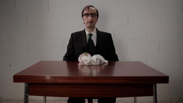 A balding man with thick-framed glasses and a strangely long nose looks directly at you. He sits at a table, upon which two white-and-brown rabbits are resting.