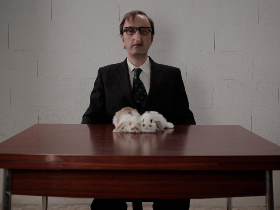 A balding man with thick-framed glasses and a strangely long nose looks directly at you. He sits at a table, upon which two white-and-brown rabbits are resting.