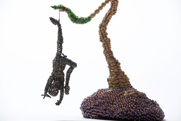 A beaded sculpture of a person made of black beads hanging upside down by a rope tied to their left foot from the branch of a tree