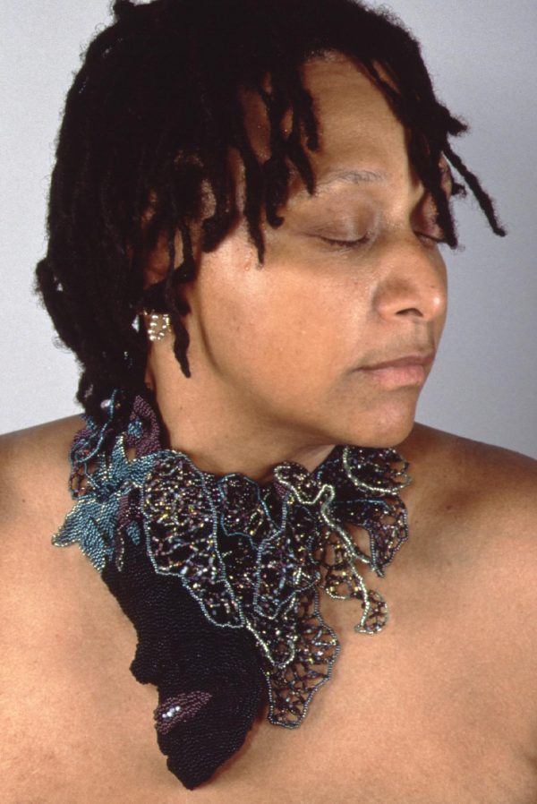 Person with bronze skin wearing a spiky necklace. Their eyes are closed as they turn towards their left.
