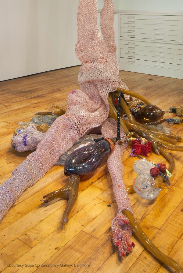 Knitted female figure with pale pink skin that has been gutted and various objects are spilling onto the wooden floor around her.