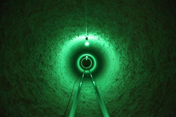 What appears to be a set of rail tracks extends straight ahead of you down a narrow tunnel. Three stark light bulbs extend downward and emit an eerie green glow.