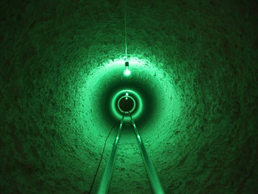 What appears to be a set of rail tracks extends straight ahead of you down a narrow tunnel. Three stark light bulbs extend downward and emit an eerie green glow.