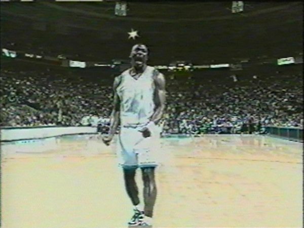 A dark-skinned basketball player wearing a light-blue uniform shouts in the middle of an empty basketball court.