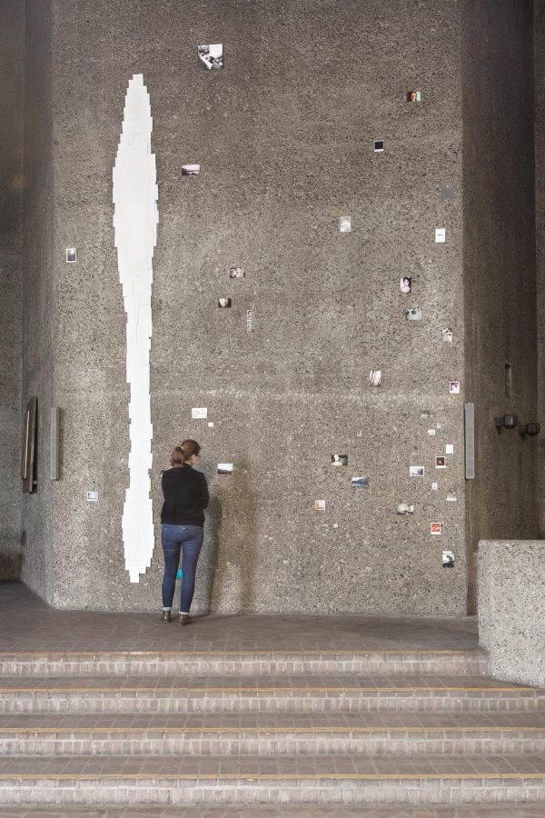 A person stands at the top of the steps next to a grey concrete wall posted with small photographs