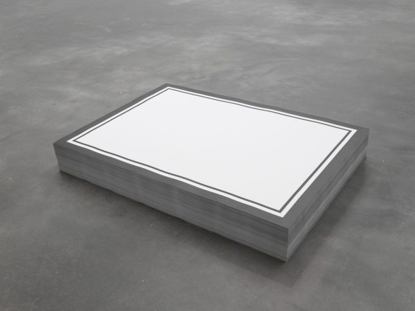 A short stack of rectangular paper on a gray concrete floor. The top piece of paper is printed with two black outlines around a solid white rectangle.