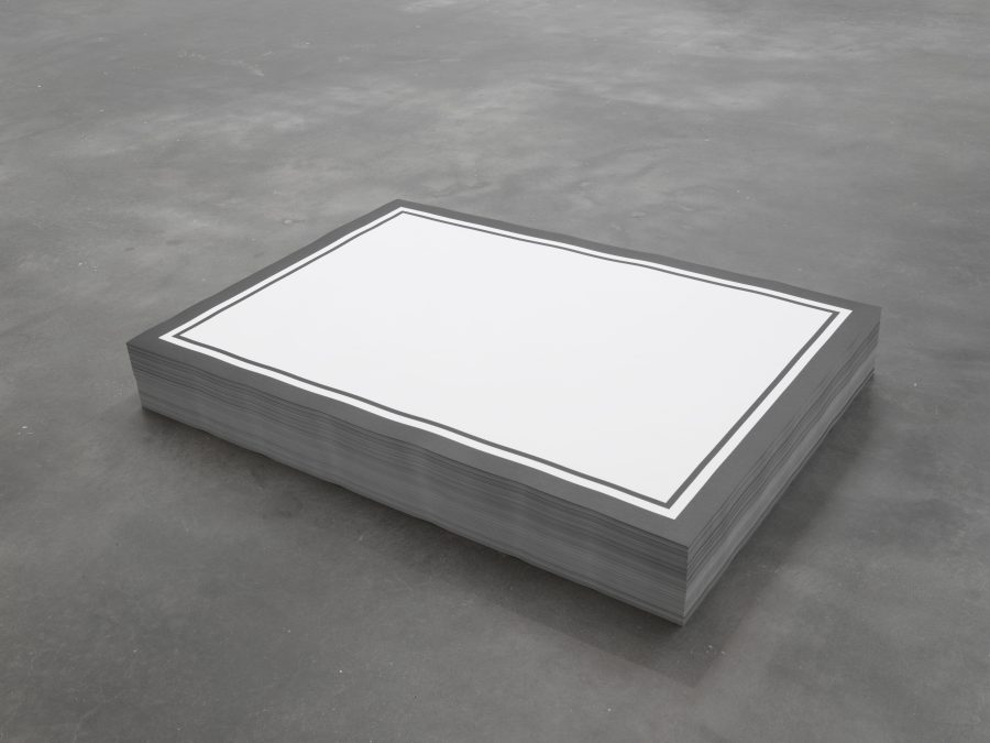 A short stack of rectangular paper on a gray concrete floor. The top piece of paper is printed with two black outlines around a solid white rectangle.