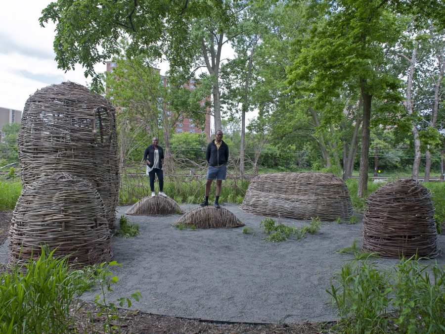 Two people stand on small mounds that are woven like wicker baskets. They are surrounded by four other similar mounds and urban greenery.