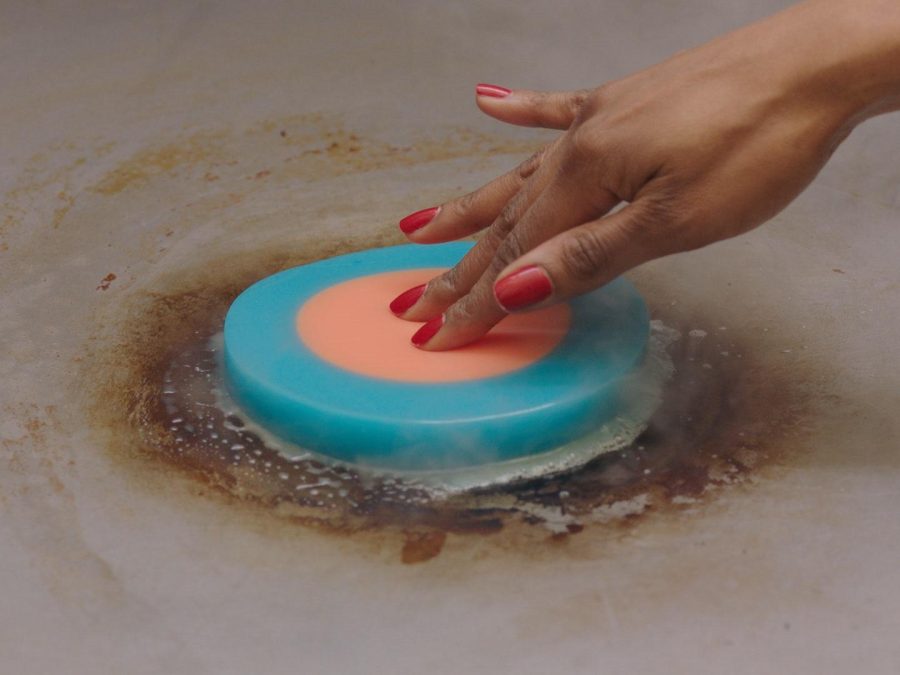A hand with red nail polish pushes down on a bright circle with a pink middle and a teal outer ring. The object has steam coming from under it.