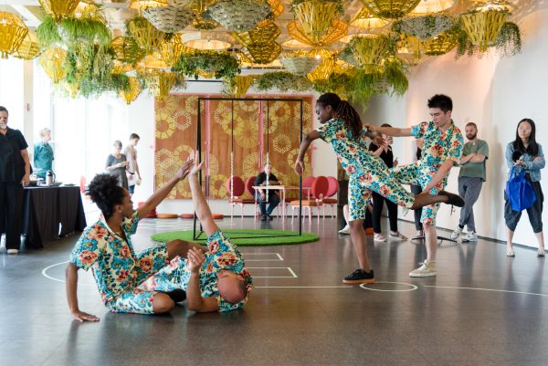 Dancers in teal patterned jumpsuits are paired in groups of two. One group is entangled on the floor; the other supports each other as the dancer on the left leans away