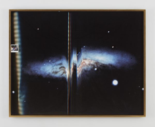 A framed artwork featuring a photograph of a galaxy with what appears to be a printing error in the middle. Inserted into the left side of the frame is a small photo of people.