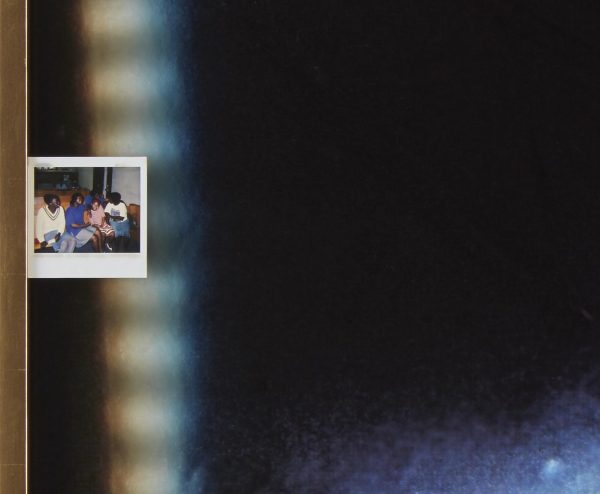 A Polaroid of four people with dark brown skin on a couch against a mostly black background aside from a vertical rainbow of light