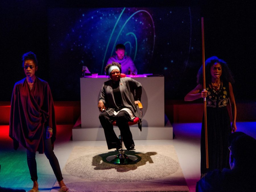 Two people, one holding a staff, stand as if guarding another person sitting in an office chair. Behind them, a person is behind a white desk lit by a purple light.