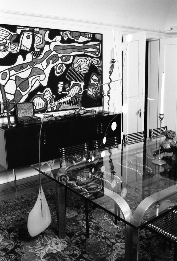 Black-and-white photo of artworks in a dining room, including a floor mobile that arches up and over a glass dining table
