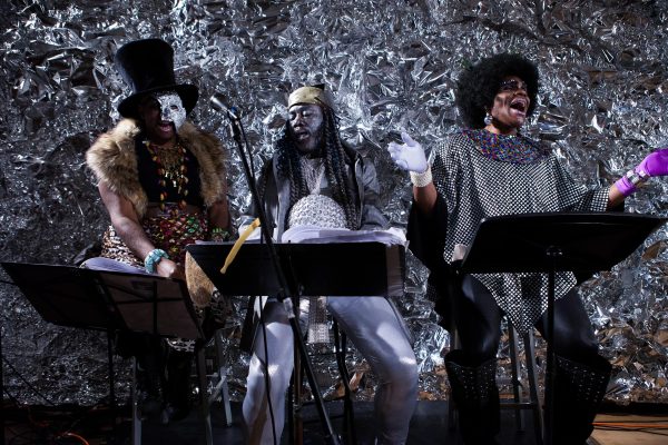 Three people, two wearing body paint, sit with music stands in front of them, mouths open to suggest they are singing