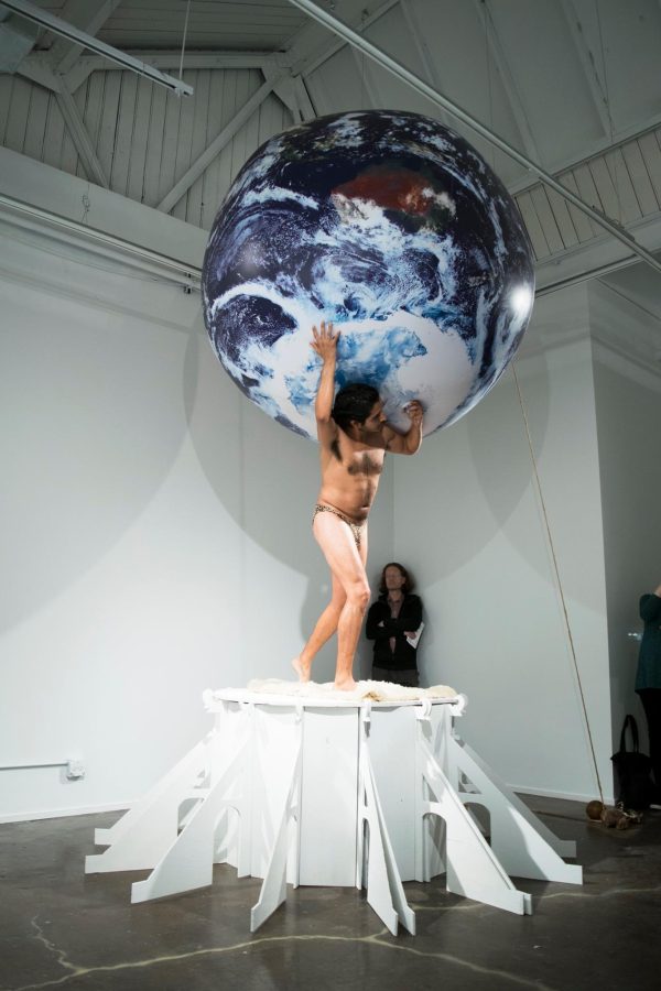 A person stands on a white pedestal holding a large Earth in a gallery space with a guard watching on