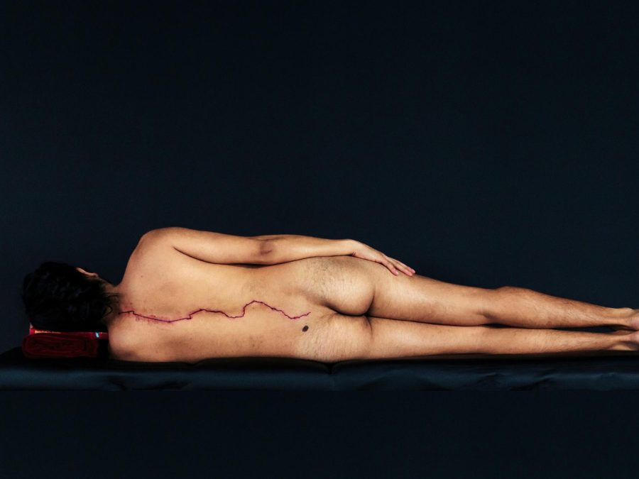 A nude person lays down on an otherwise black setting, their back facing us to display a large red line that traces the spine