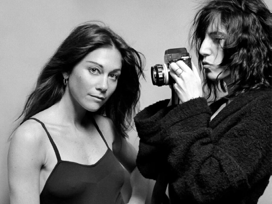 Black and white photo of two people dressed in dark clothing. The person on the right holds up a camera to the other, who looks directly at the viewer.