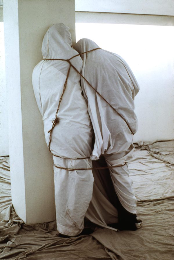 Two people wrapped in white canvas and rope stand close together against a white column in a room with canvas covering the floor as well