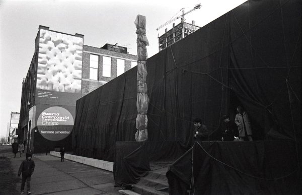 Black and white image of a building that has been wrapped in a dark material, and an identifying sign attached to the building that has been wrapped in a light fabric. Behind the building a mural features a painting of a pastry, and a sign reading Museum of Contemporary Art, Current Exhibitions, Christo, Westermann, become a member.