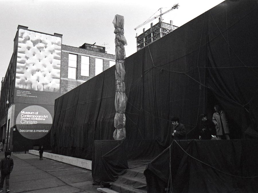 Black and white image of a building that has been wrapped in a dark material, and an identifying sign attached to the building that has been wrapped in a light fabric. Behind the building a mural features a painting of a pastry, and a sign reading Museum of Contemporary Art, Current Exhibitions, Christo, Westermann, become a member.