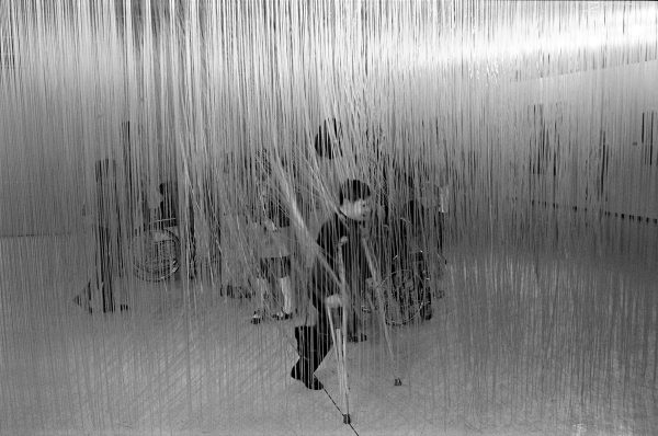 A group of children, some pushed in wheelchairs or using crutches, touches strands of an artwork hanging from the ceiling.