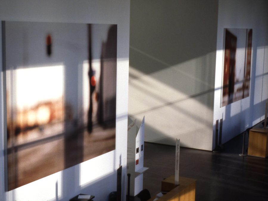 Two large, blurry images of streets hang in a two-story space lit dramatically by long shadows cast by sunlight