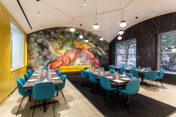 Teal chairs line three long tables in a room with a colorful mural at the back.