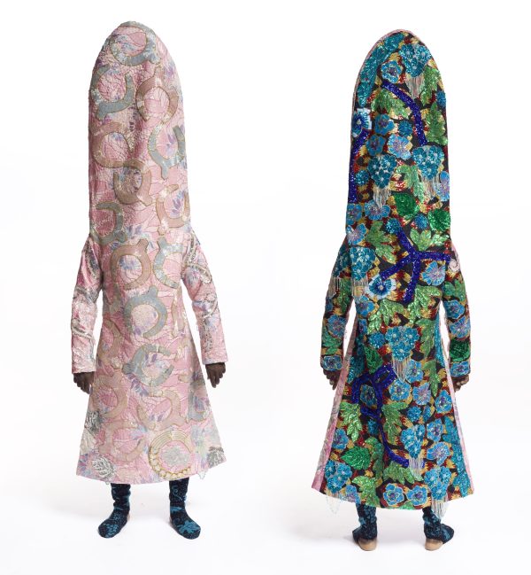 Two humanoid figures wearing elaborate costumes stand, one facing you and one facing away. One wears a light pink costume and the other wears a vivid green, purple, and blue costume.