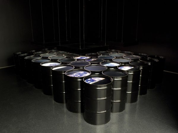Rows of black barrels in a v formation filled with liquid in a dark room painted black. Images are projected onto the surface of some barrels.