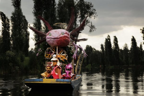 A group of people on a flat-bottomed boat with a giant winged, salamander-like creature attached above them in a tropical setting