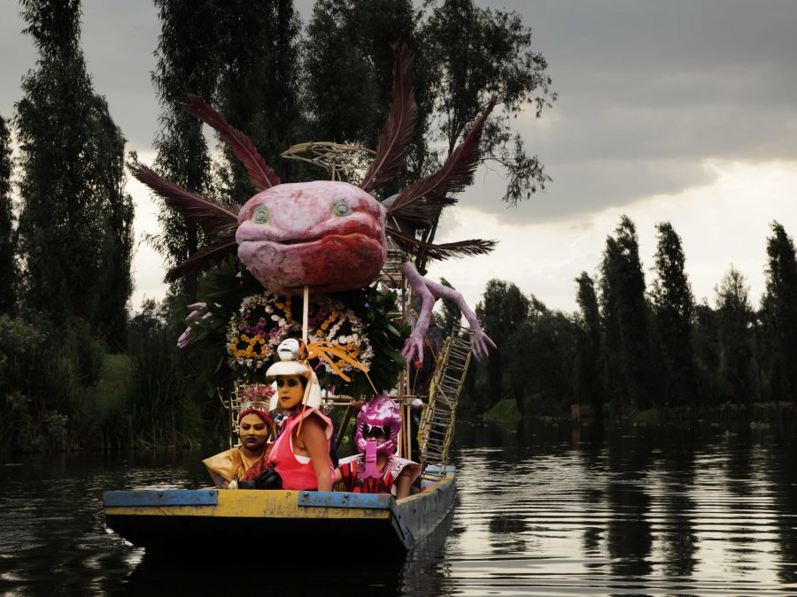 A group of people on a flat-bottomed boat with a giant winged, salamander-like creature attached above them in a tropical setting