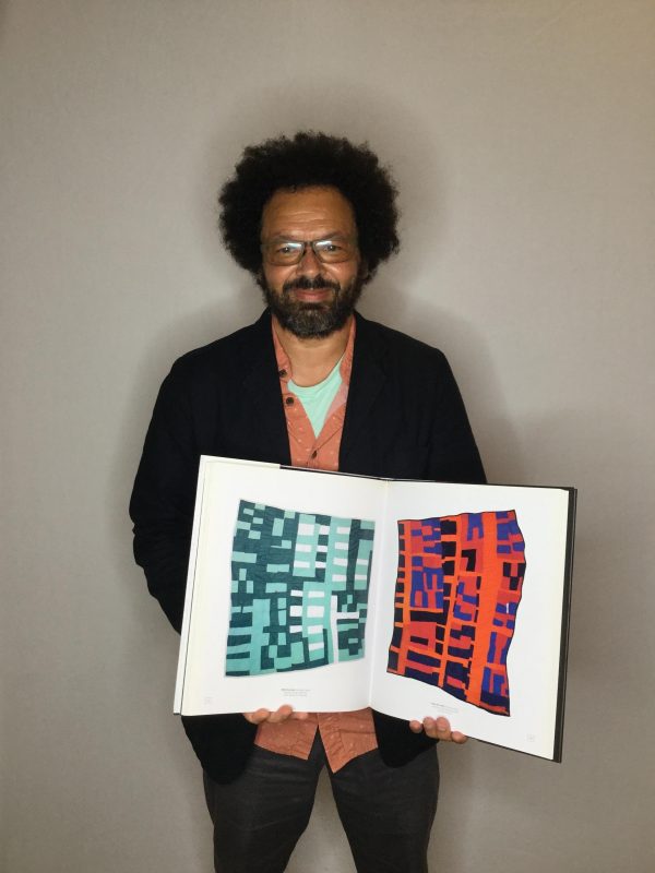A male-presenting person with curly black hair and a tightly shaved beard poses with a book opened to two artworks, one teal and one red